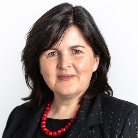 a woman with dark shoulder length hair, friendly smile, wearing a dark suit and red bead necklace