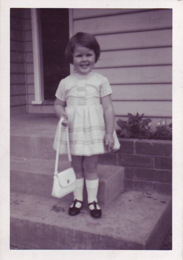 black and white photo of a smiling four year old girl with short dark hair, wearing a party dress, white socks and black shoes and holding up a white handbag with a long strap. She is standing on the bottom concrete step at the from of her weatherboard house.